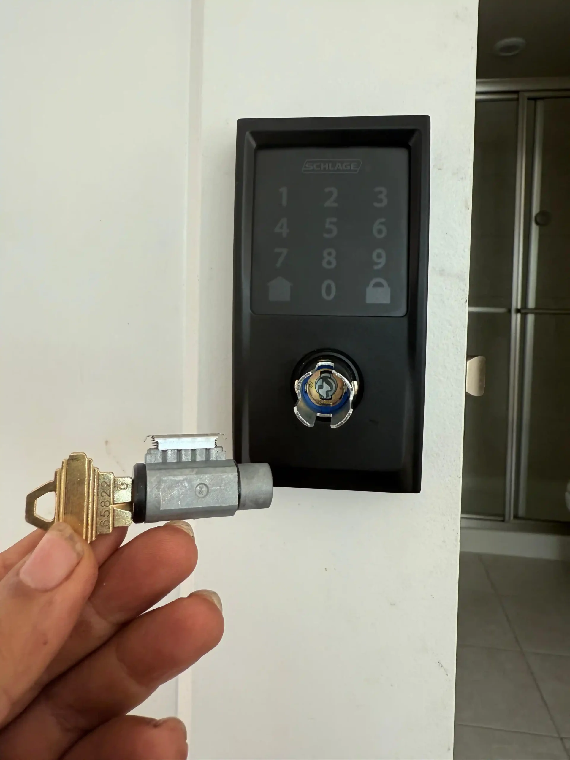 A hand holds a key and lock cylinder in front of a modern black keypad deadbolt by Schlage, demonstrating AA Locksmith Near Me LLC's residential lock rekeying service.